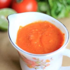 Momos Tomato Chutney on a white floural printed cup on a creamy coloured surface and some lemon,tomato and some coriander leaves on the background|