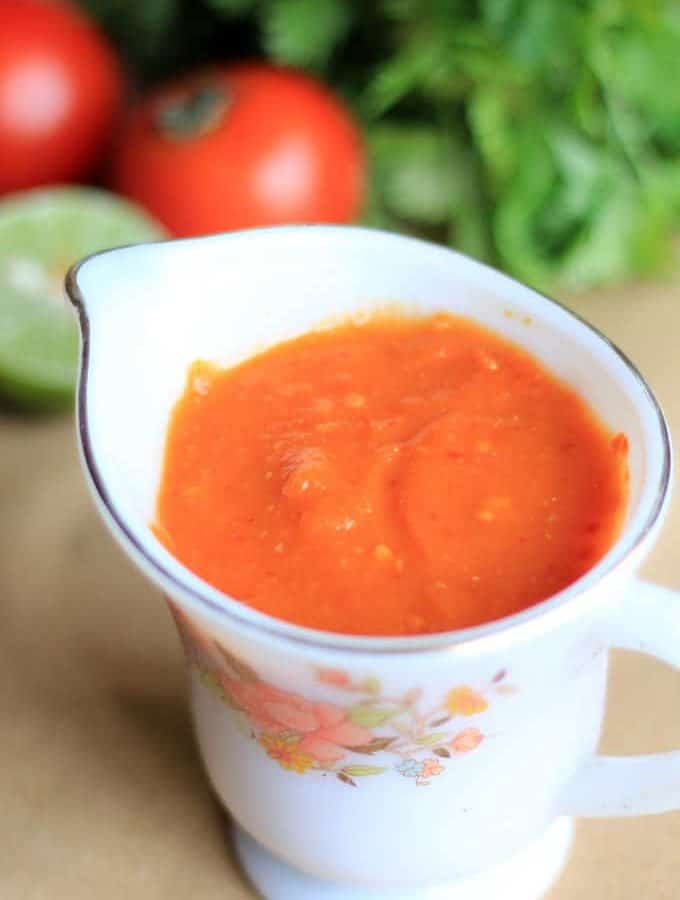 Momos Tomato Chutney on a white floural printed cup on a creamy coloured surface and some lemon,tomato and some coriander leaves on the background|