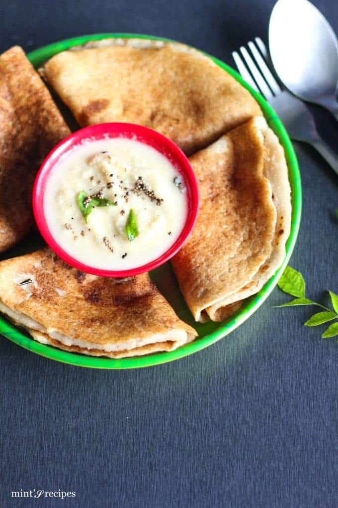 Oats Poha Dosa on a round plate with 4 pieces of oats dosa on it with a small bowl of coconut chutney and little tempering of mustard seed and curry leaves putted on a dark surface with spoon and fork and some curry leafs |