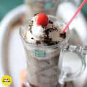 Oreo-Milk-Shake on a white surface with 2 glasses of milkshake decorated with some heavy cream with some oreo biscuit crush and a cherry on the top with a strow in it |