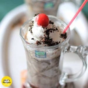 Oreo-Milk-Shake on a white surface with 2 glasses of milkshake decorated with some heavy cream with some oreo biscuit crush and a cherry on the top with a strow in it |