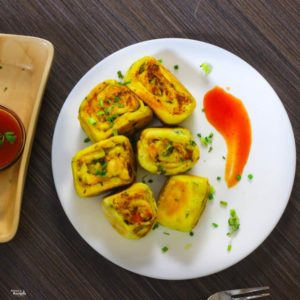 Paneer Bhurji Swiss Roll on a white plate with some swiss rolls and tomato ketchup spread on one side, garnished with spring onion and a small bowl of tomato ketchup on the background