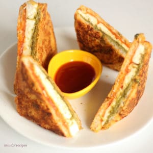 Bread Paneer Pakoda on a white plate with some tomato ketchup and green chutney on a small bowl kept on a dark mattress