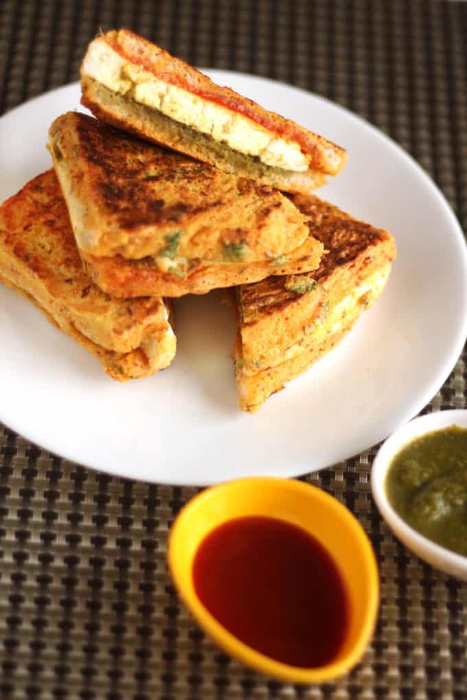 Bread Paneer Pakoda on a white plate with some tomato ketchup and green chutney on a small bowl kept on a dark mattress 