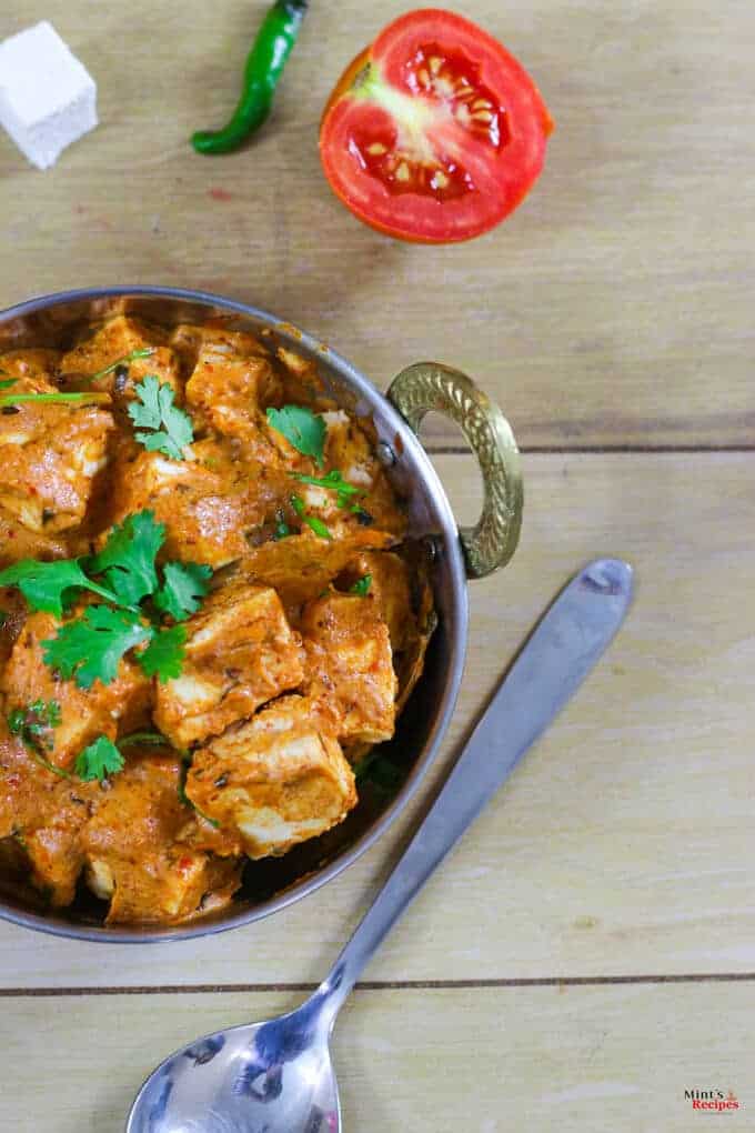 Paneer Makhni on a deep vessel kept on a wooden surface with garnishing of coriander leaves and a spoon kept beside it and some tomato slices, chili and paneer cubes on the background |