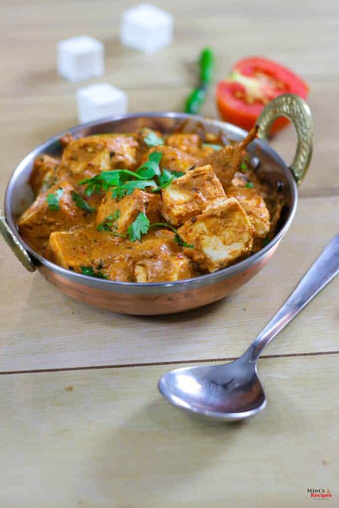 Paneer Makhni on a deep vessel kept on a wooden surface with garnishing of coriander leaves and a spoon kept beside it and some tomato slices, chili and paneer cubes on the background |