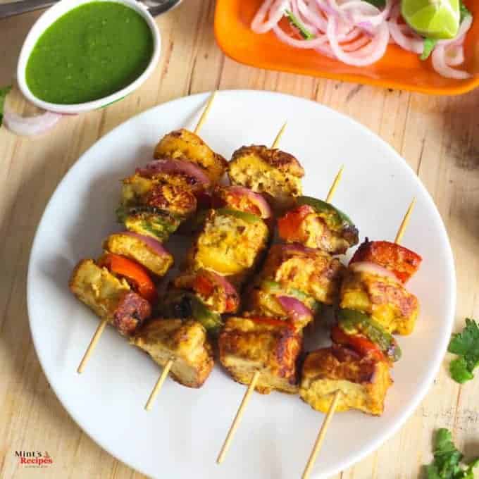 Paneer Tikka on a white plate with some green chili ,lemon and onion rings with some green chutney 