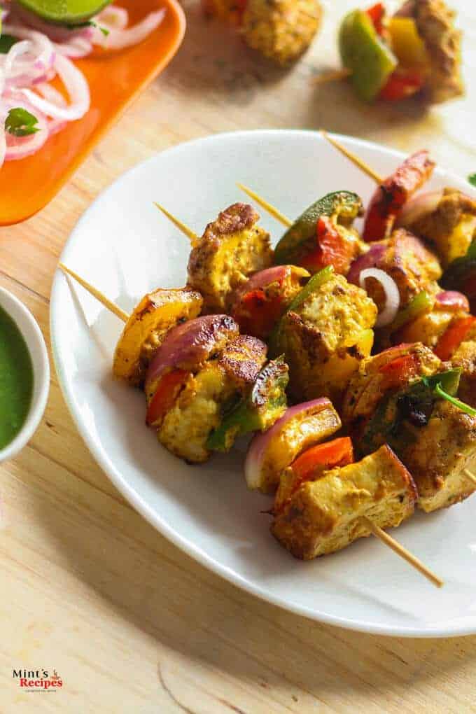 Paneer Tikka on a white plate with some green chili ,lemon and onion rings with some green chutney 