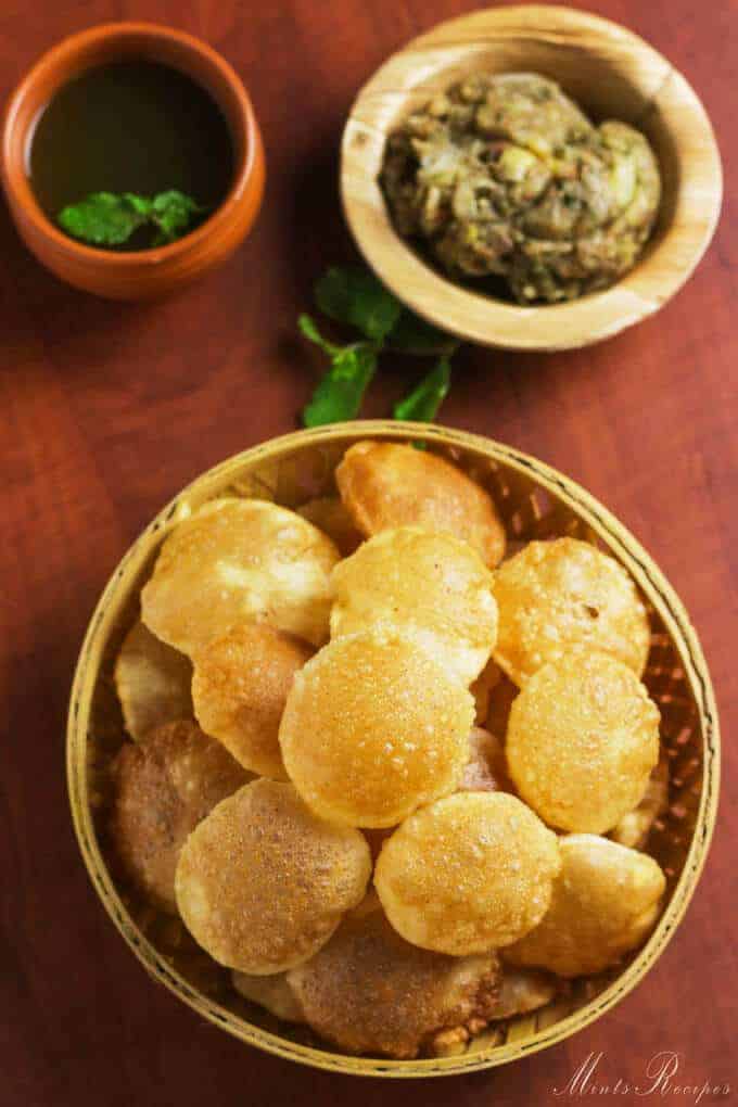 Pani Puri on a wooden basket full of puri's and potato mixture on a wooden plate and some sour water in a clay pot with some coriander leaves in it |