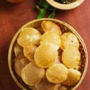 Pani Puri on a wooden basket full of puri's and potato mixture on a wooden plate and some sour water in a clay pot with some coriander leaves in it |