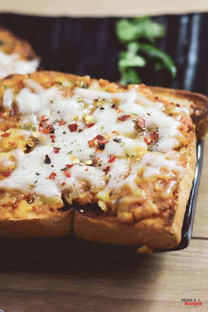 Potato cheese toast on a black plate with some potato mixture and grated cheese with some italian seasoning and red chilli flakes| www.mintsrecipes.com |