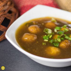 Potato Manchurian On a white bowl with some spring onions on top for garnishing and a wooden bowl with some mat on the background |