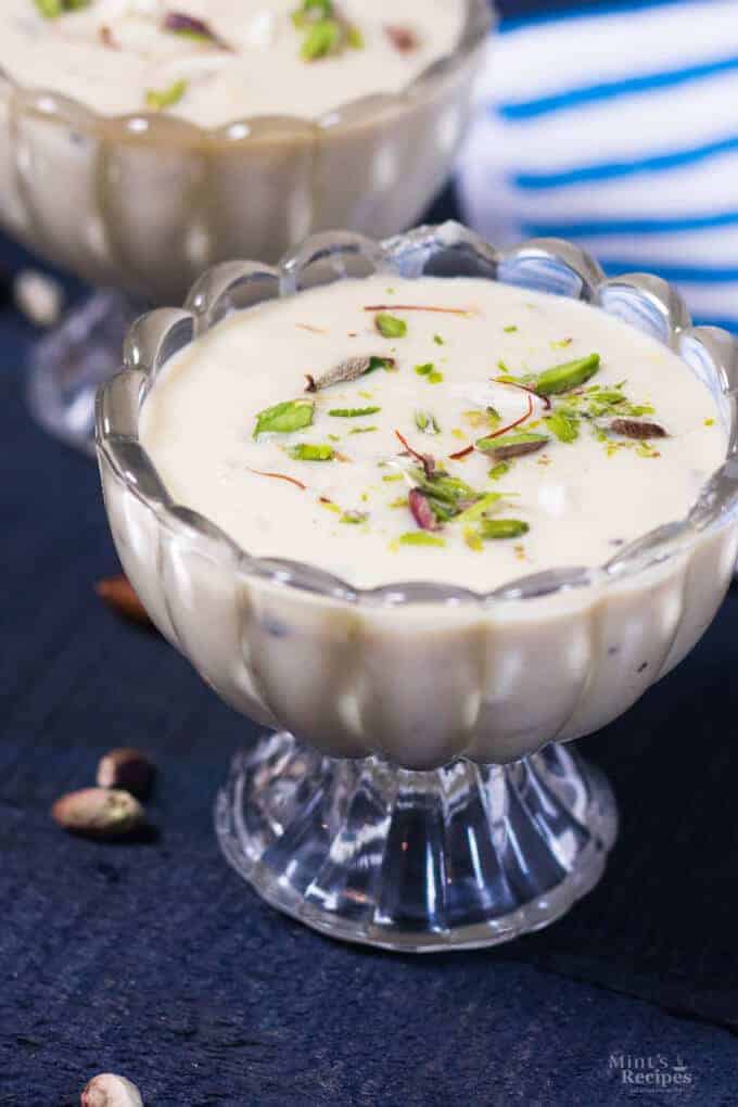 Rabdi on a transparent bowl full of rabdi on it with some pista and few string of saffron on it. kept on a wooden dark surface with some pista spread on it with another full bowl of rabdi on the background |