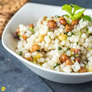 Sabudana ki khichri on a white bowl with some coriander leaves on it and some nuts spread on a wooden surface and a basket behind the bowl |