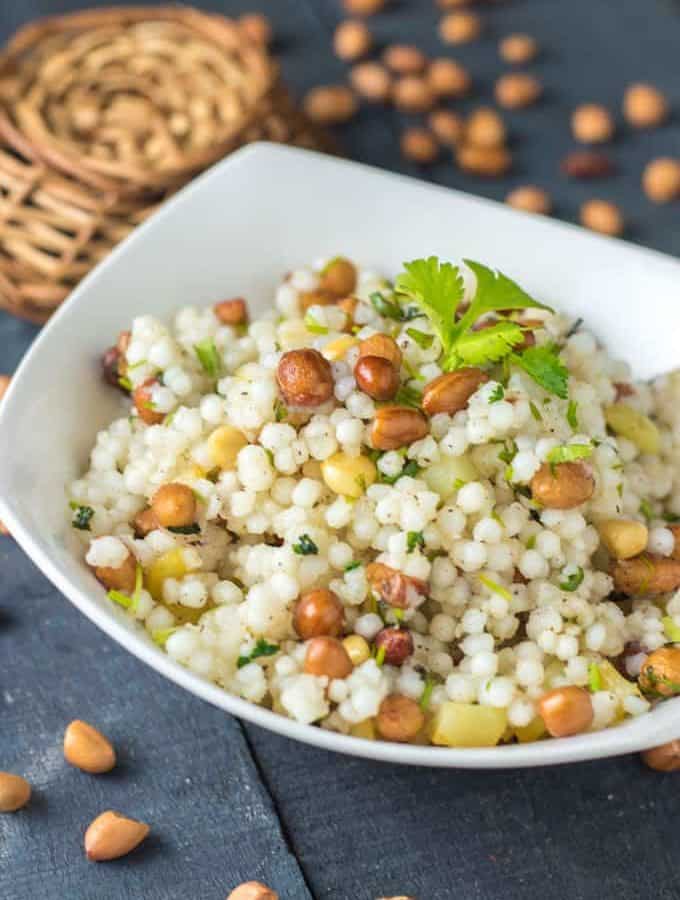 Sabudana ki khichri on a white bowl with some coriander leaves on it and some nuts spread on a wooden surface and a basket behind the bowl |