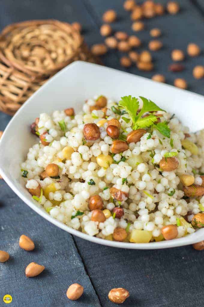 Sabudana ki khichri on a white bowl with some coriander leaves on it and some nuts spread on a wooden surface and a basket behind the bowl |