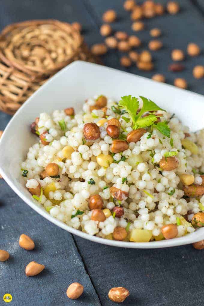 Sabudana ki khichri on a white bowl with some coriander leaves on it and some nuts spread on a wooden surface and a basket behind the bowl 