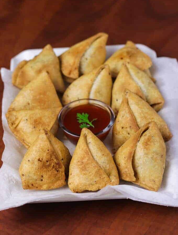 Samosa on a white tray with some tissue paper and little tomato ketchup on the center of the plate with rounded into samosas kept on a dark wooden surface |