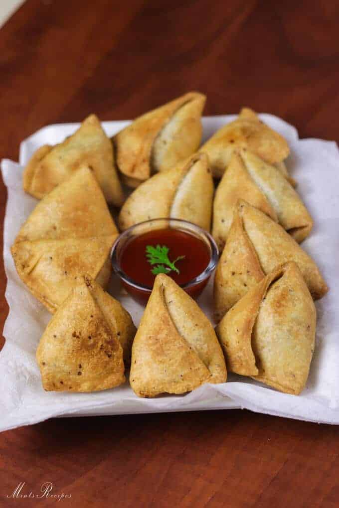 Samosa on a white tray with some tissue paper and little tomato ketchup on the center of the plate with rounded into samosas kept on a dark wooden surface |