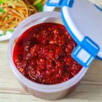 Schezwan Sauce on a airtight container with a plate of noodles on it kept on a wooden surface |