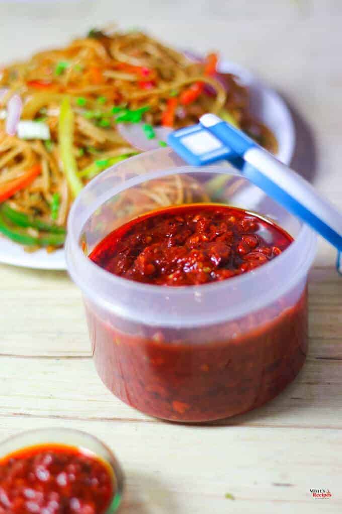 Schezwan Sauce on a airtight container with a plate of noodles on it kept on a wooden surface |
