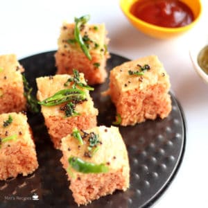 Suji Dhokla on a black plate tempered with a white background and some tomato sauce on a yellow small bowl