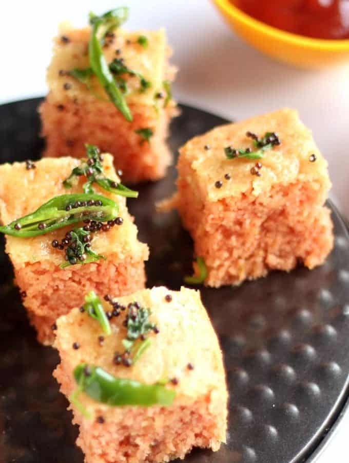 Suji Dhokla on a black plate with tempering of green chili and mustard seed |
