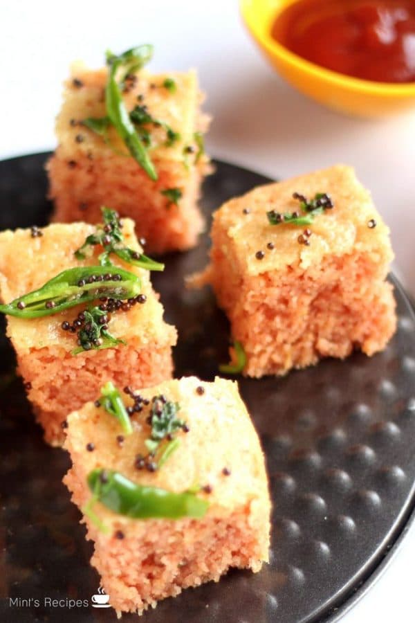 Suji Dhokla on a black plate with some tempering of green chili and mustard seed with some tomato ketchup on the background