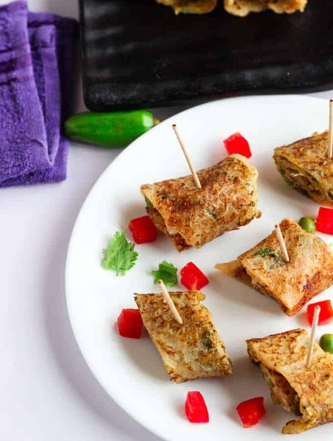 Suji Potato Rolls on a white plate with some chopped veggies like red bell pepper and green peas with some coriander leaves and some suji potato rolls in a black tray kept on a white surface |
