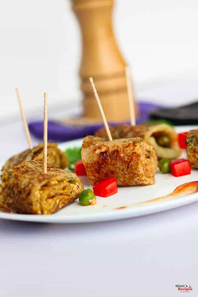 Suji Potato Rolls on a white plate with some chopped veggies like red bell pepper and green peas with some coriander leaves and some suji potato rolls in a black tray kept on a white surface |