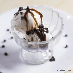 Vanilla Ice Cream on a glass ice cream bowl whit 2 scoops of vanilla ice cream and spreaded some chocolate syrup and choco chips on it kept on a white plate |