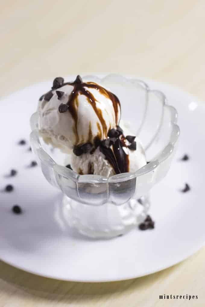 Vanilla Ice Cream on a glass ice cream bowl whit 2 scoops of vanilla ice cream and spreaded some chocolate syrup and choco chips on it kept on a white plate |
