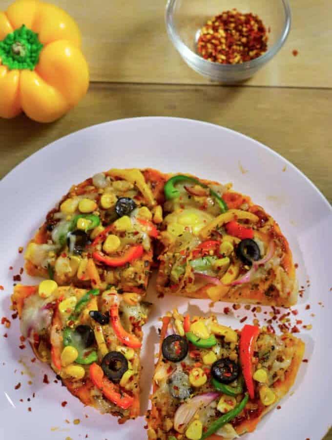 Veg Pizza On Tawa on a white plate with some sprinkles of chilli flakes kept on a wooden surface with a yellow bell pepper and a small bowl of red chilli flakes on the background |