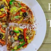 Veg Pizza On Tawa on a white plate with some sprinkles of chilli flakes kept on a wooden surface with a yellow bell pepper and a small bowl of red chilli flakes on the background |