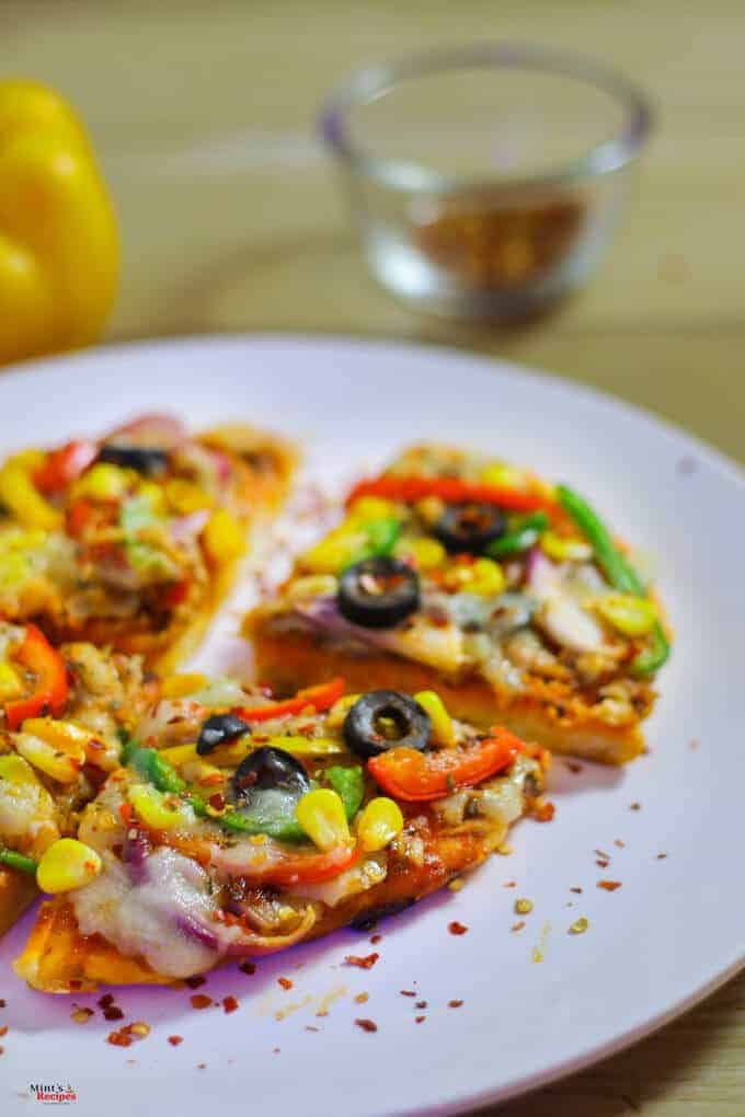 Veg Pizza On Tawa on a white plate with some sprinkles of chilli flakes kept on a wooden surface with a yellow bell pepper and a small bowl of red chilli flakes on the background |
