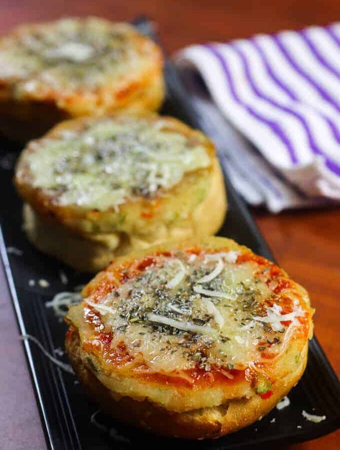 Veg Suji Buns on a black tray with some suji buns garnished with some cheese and oregano kept on a wooden surface |