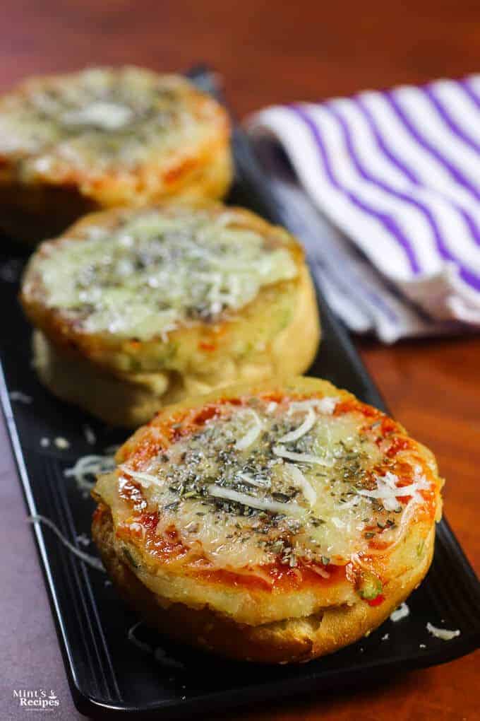 Veg Suji Buns on a black tray with some suji buns garnished with some cheese and oregano kept on a wooden surface |